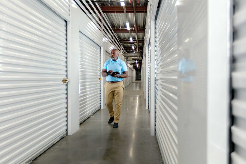 A man working at a storage unit is reviewing information on a tablet while walking down a hall surrounded by units.