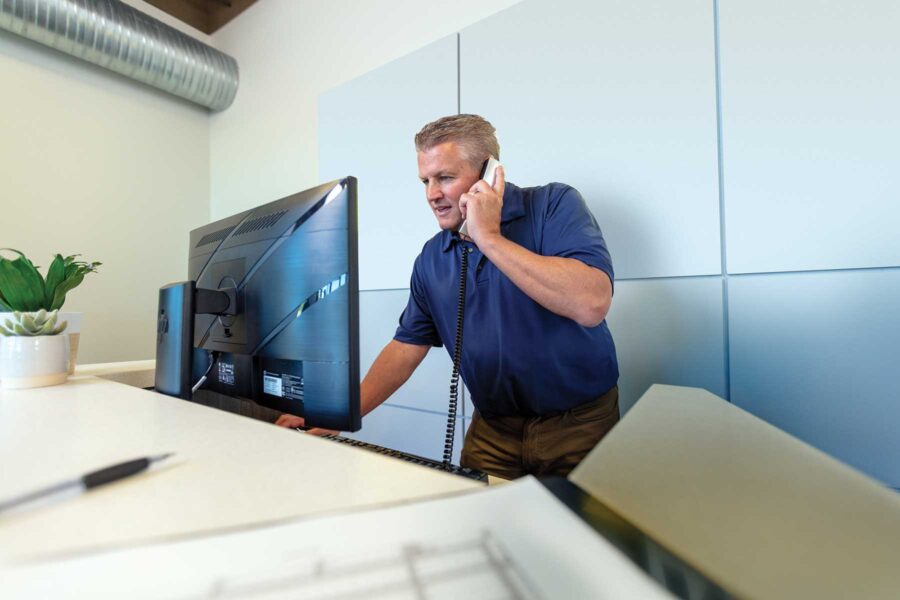 A man in a blue polo is on the phone with a customer and reviewing his computer.